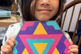 Eva is holding up a colorful star mosaic pattern that she created. 