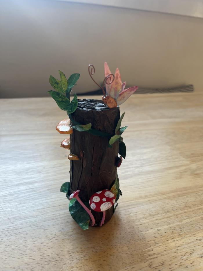 A toilet paper roll covered in tissue paper and painted and red hot glue mushrooms on the side, at watercolor butterfly on top, a leather and paper vine wrapped around and some hot glue ladybugs